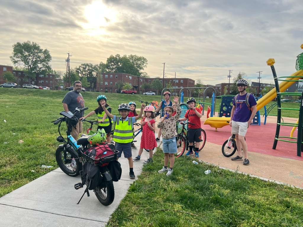 About a dozen children in helmets and hi-vis vests, with adults and bikes, gathered on the path beside the playground at Kirkwood Neighborhood Park.