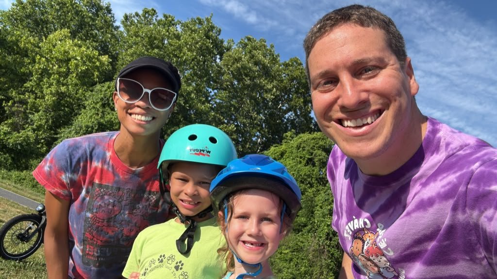 Two adults and two children in bike helmets, smiling on a sunny Friday morning ride.